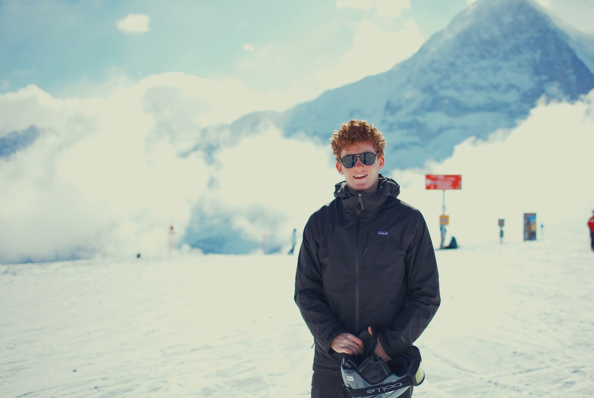 Ryan McComb standing on a snowy alpine slope in the Swiss Alps, sunglasses on, holding a ski helmet, with a snow-capped peak rising through clouds behind him